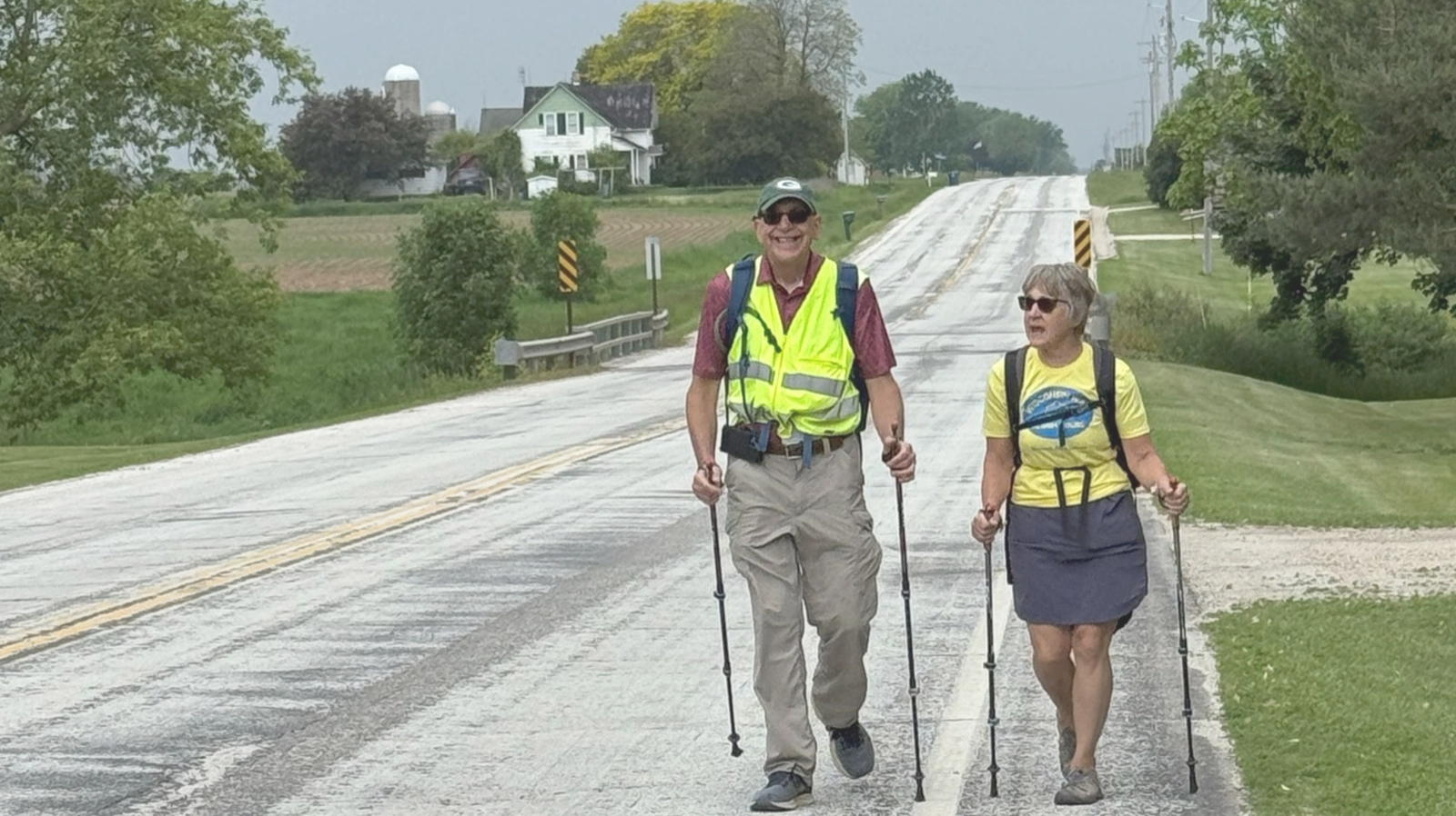 Two pilgrims in safety vests walking with trekking poles along a rural road