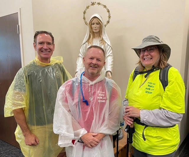 Three smiling pilgrims in rain ponchos beside a statue of Mary