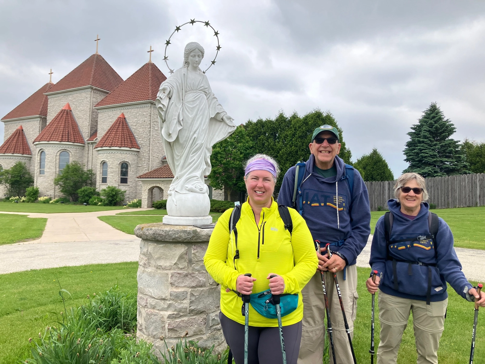 Three pilgrims with trekking poles beside a white statue of Mary in front of a church