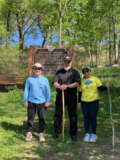 Three pilgrims, one a priest with a walking staff, beside the St. Mary of the Oaks historical marker