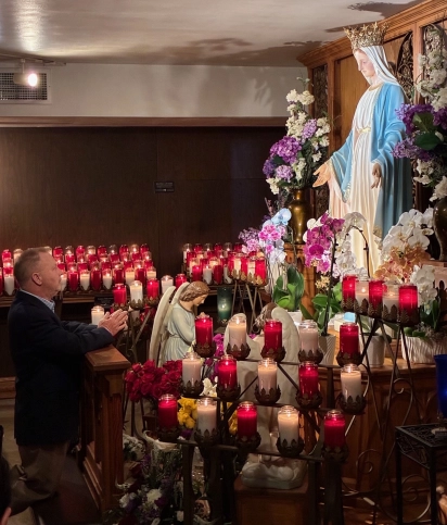 A pilgrim kneeling in prayer before a statue of Mary surrounded by red votive candles