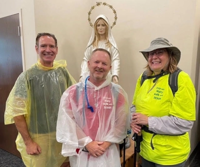 Three smiling pilgrims in rain ponchos beside a statue of Mary